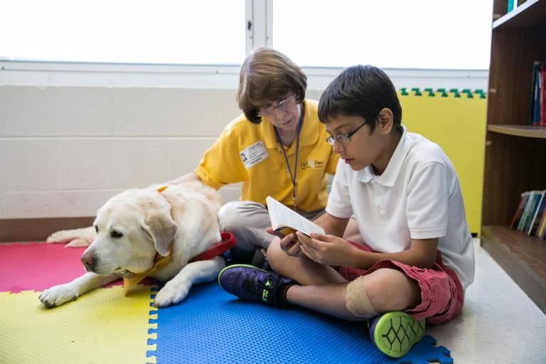 Volunteer and child read together beside a therapy dog in a classroom.