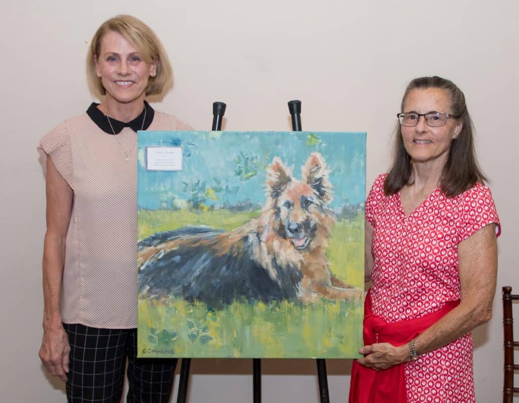 Two women display a painting of a dog at a Hand in Paw gallery party.