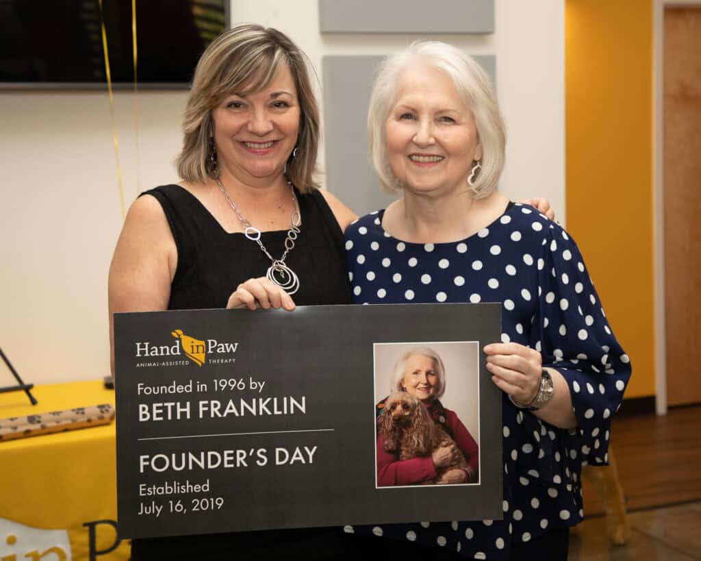 Two women hold a Hand in Paw Founder’s Day plaque honoring founder Beth Franklin.