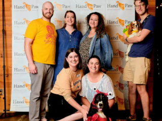 Barktoberfest 2021 Brittany Filby and others pose in front of a Hand in Paw "step and repeat" banner at Barktoberfest in 2021.