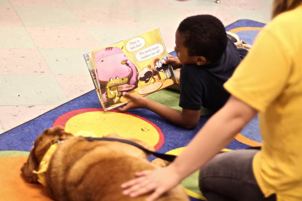Child reads beside a therapy dog and volunteer during an animal-assisted education visit.