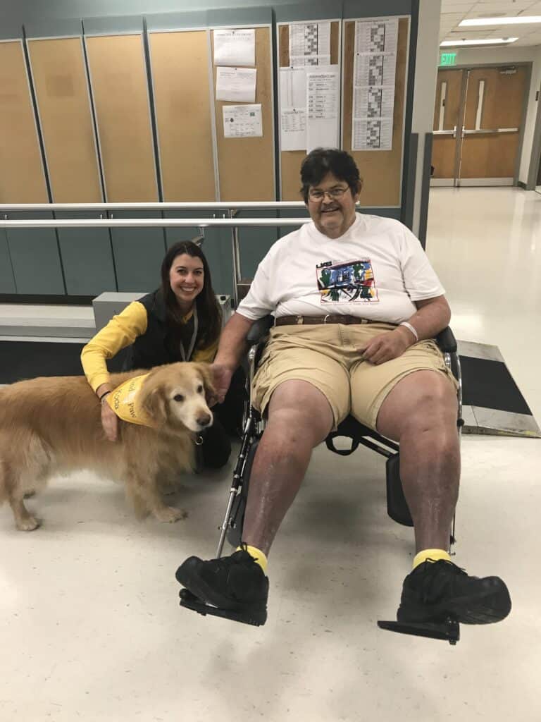 Therapy dog and volunteer visiting a person in a wheelchair at a healthcare facility.