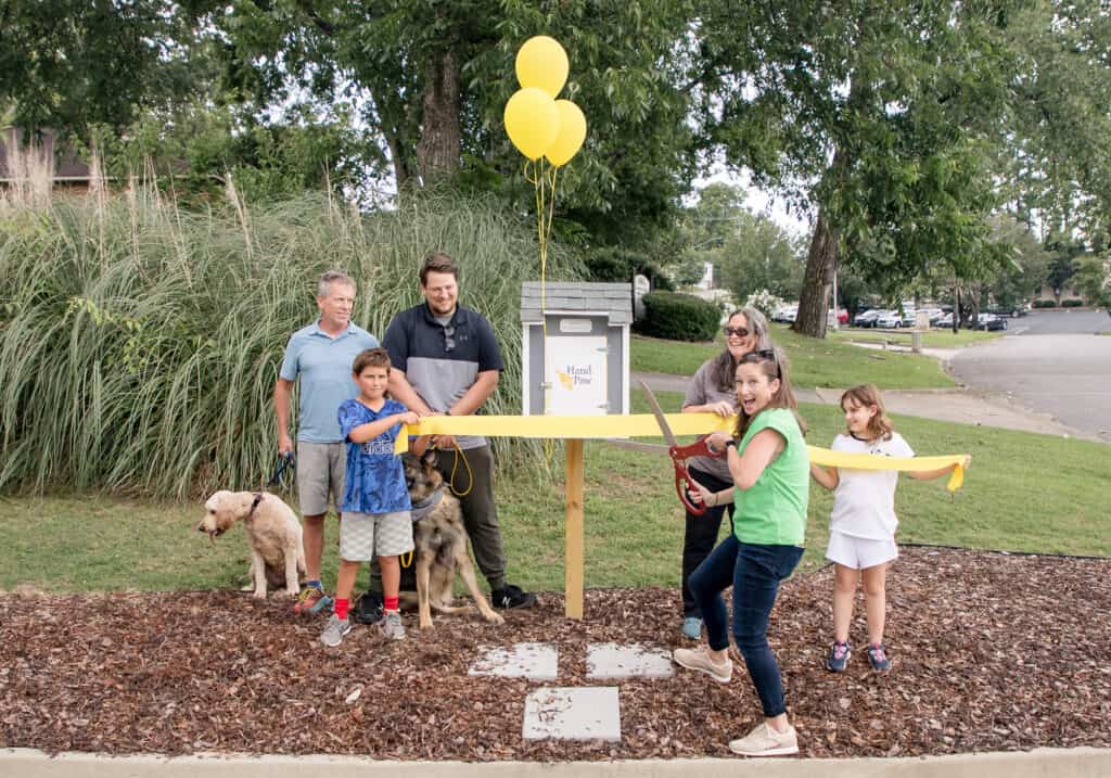Children, adults, and dogs gather outdoors beside a ribbon and balloons at a Hand in Paw event.