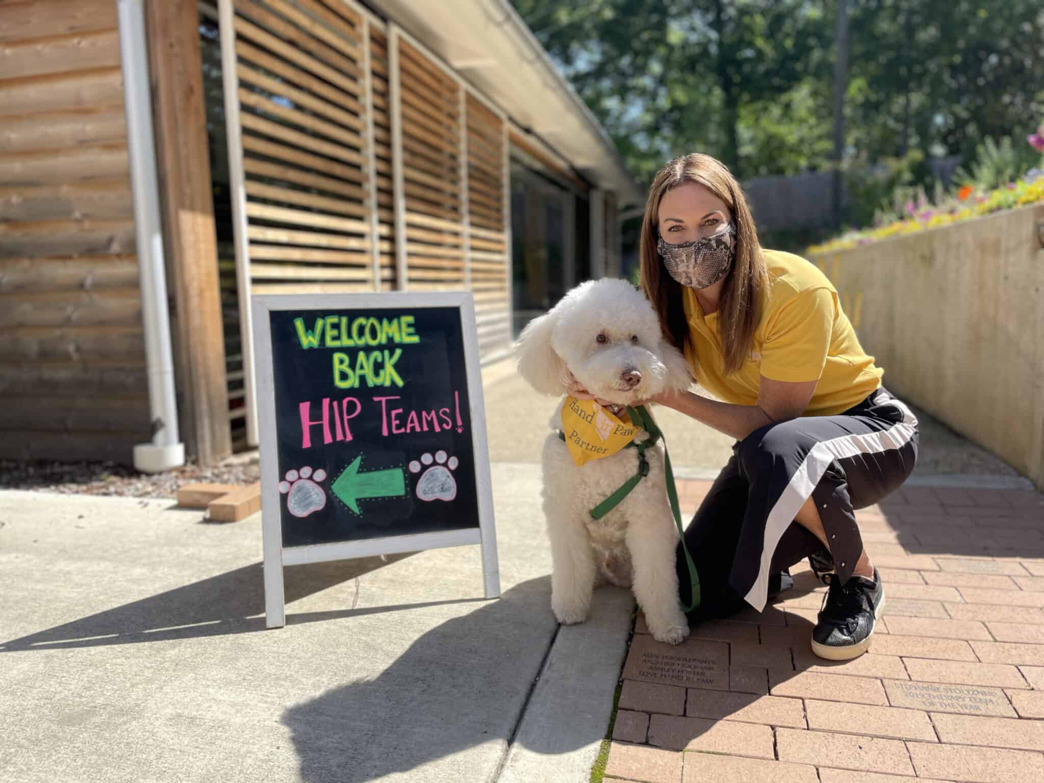 Hand in Paw therapy team member with a dog beside a welcome-back sign