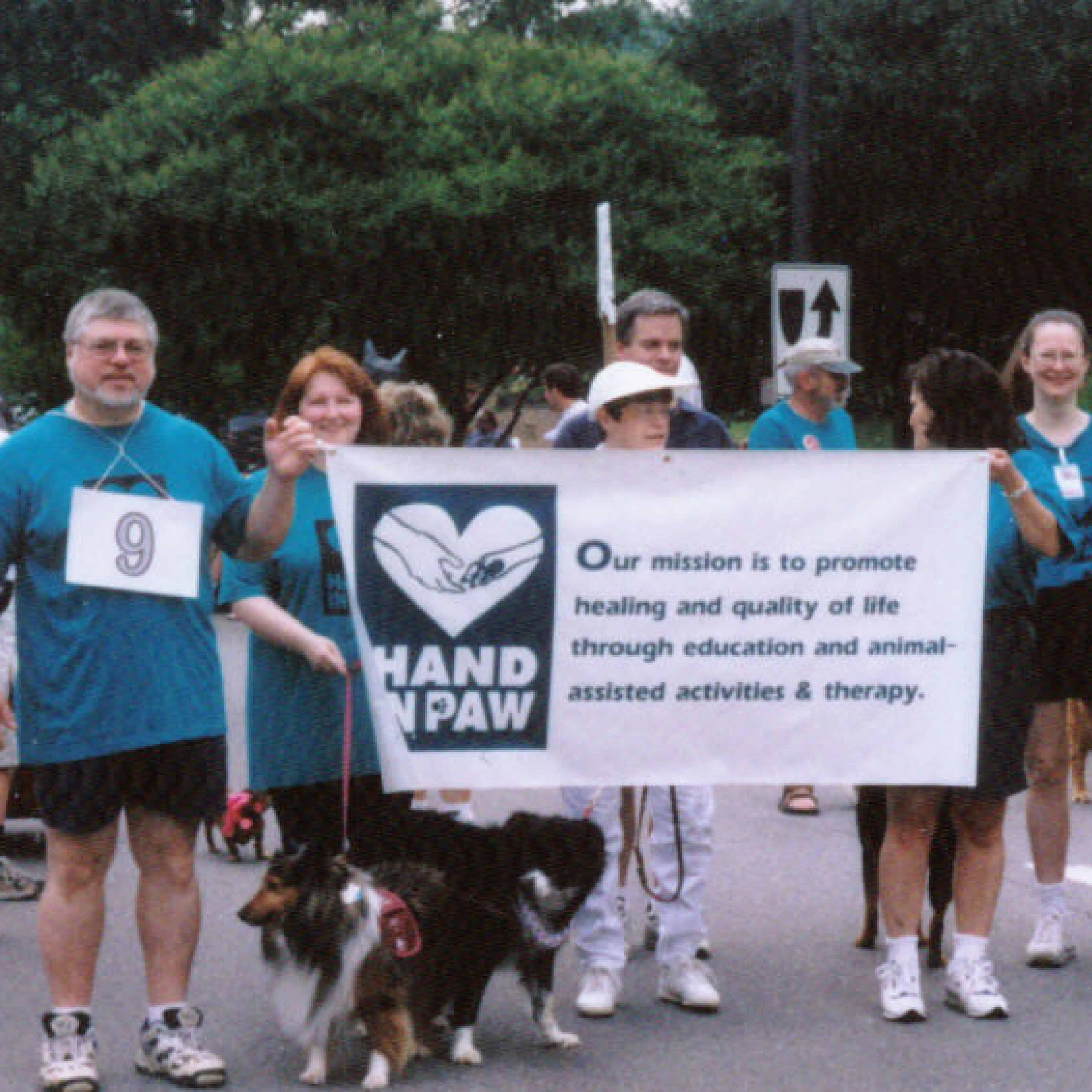 Hand in Paw volunteers and dogs carry a banner promoting animal-assisted activities and therapy.