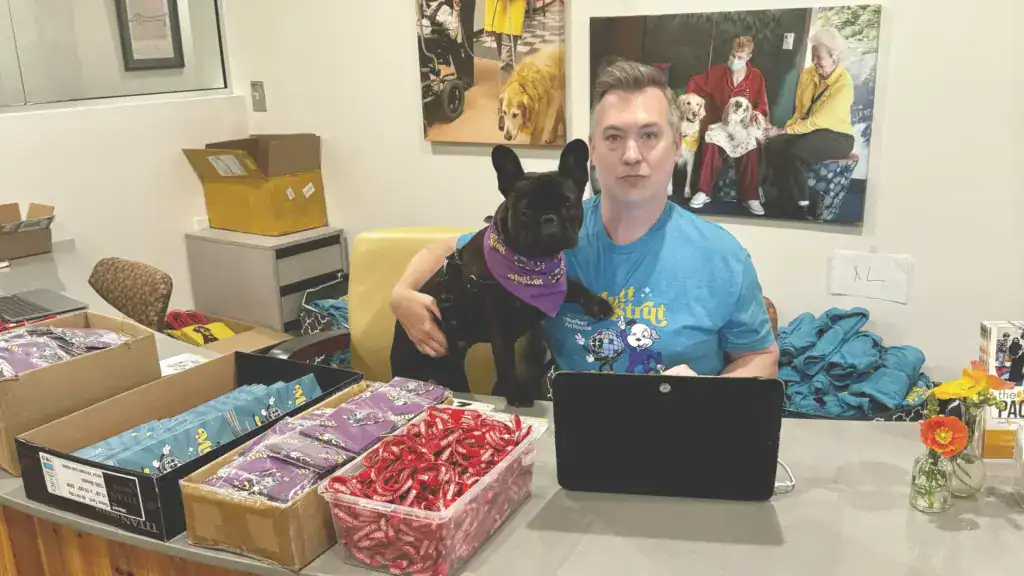 Hand in Paw volunteer holds a dog beside a laptop and boxes of outreach materials.
